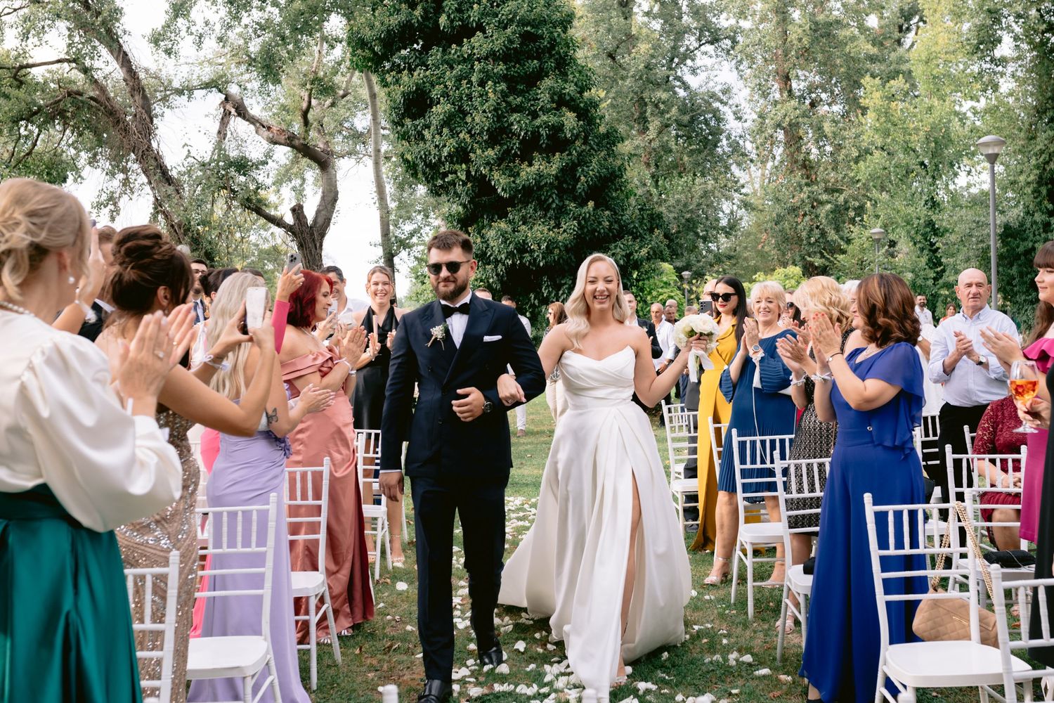 Bride and groom walk down outdoor aisle as guests applaud at a sunny garden wedding ceremony.
