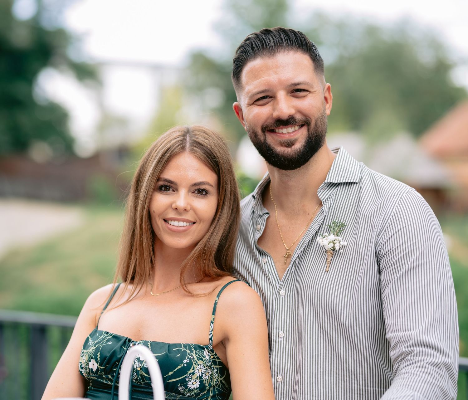 A smiling couple poses outdoors, woman in green floral dress and man in striped shirt with boutonniere.