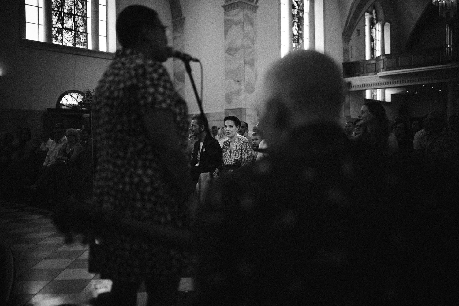 Black and white photo of a singer performing at a microphone inside a historic church with stained glass windows.