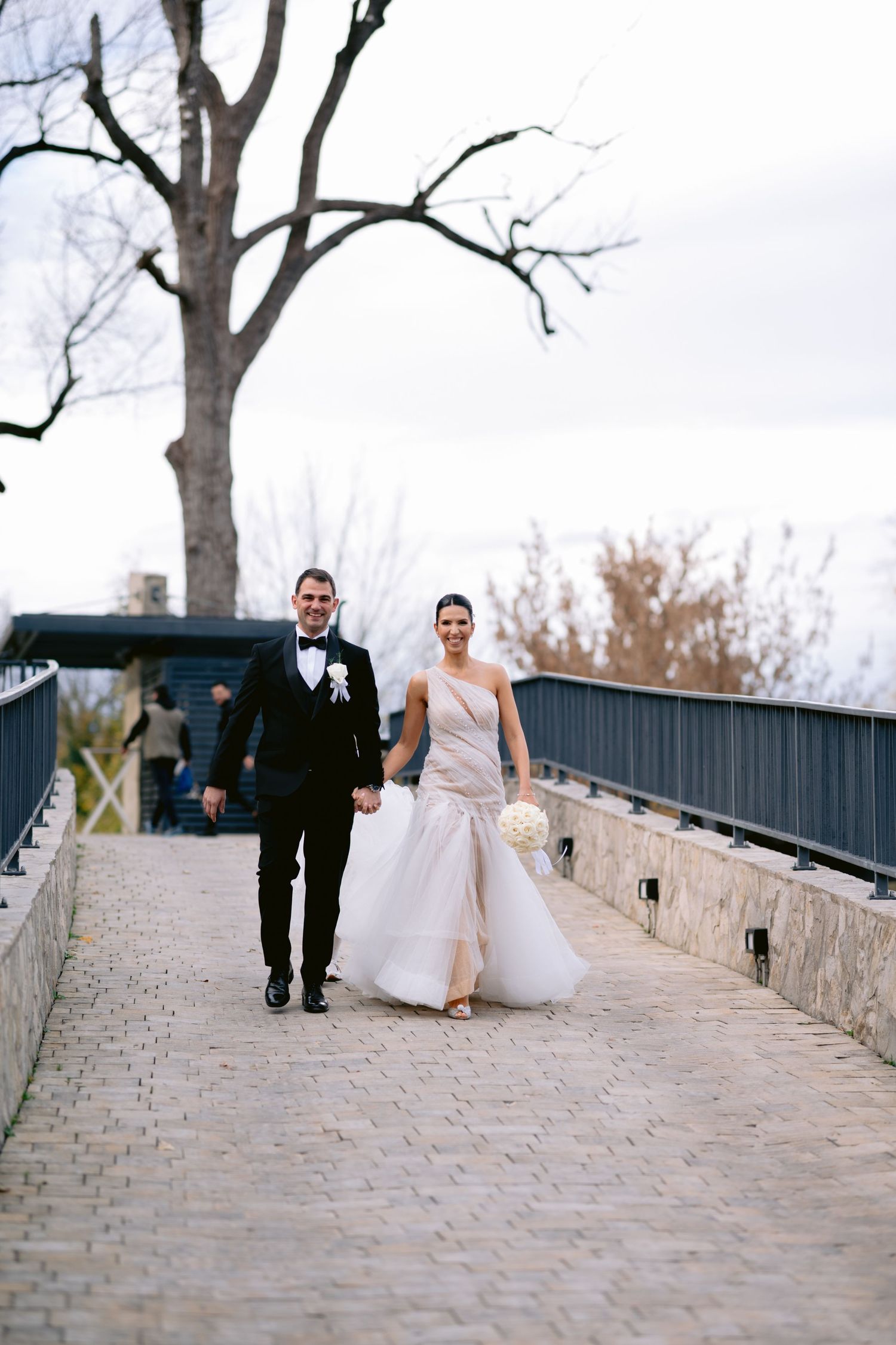 Bride and groom walk hand-in-hand on cobblestone bridge, she in white gown holding bouquet, he in black tuxedo.