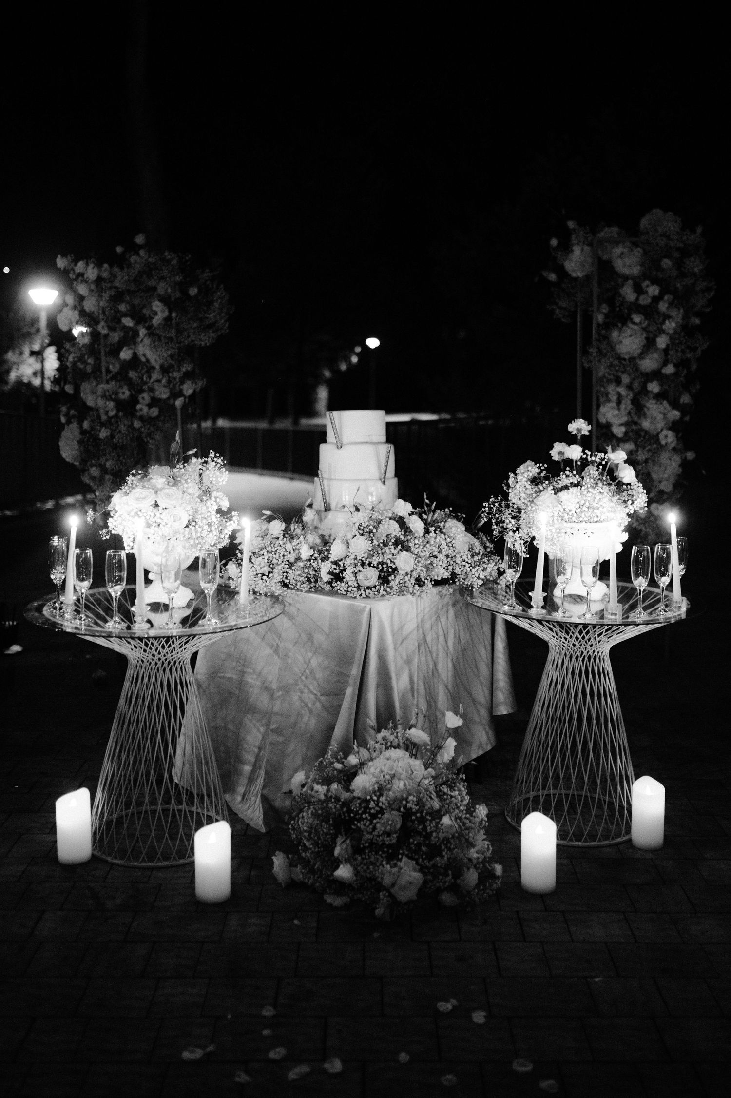 Black and white photo of elegant wedding cake table with candles, floral arrangements, and decorative wire stands at night.