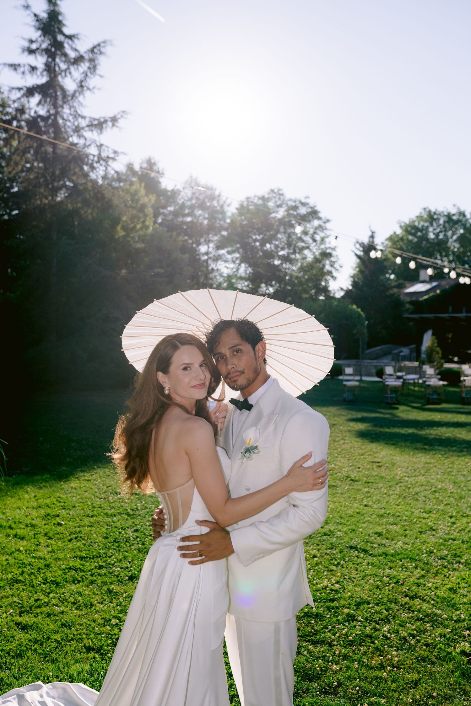 Bride and groom embrace under white parasol on sunny green lawn, groom in white suit with boutonniere, bride in elegant backless gown.