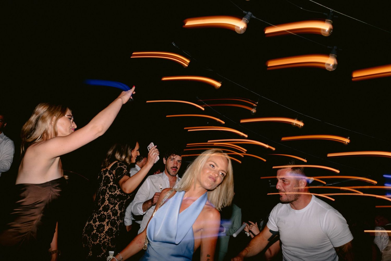 People dancing at a lively nightclub party with colorful light trails streaking across the dark background.