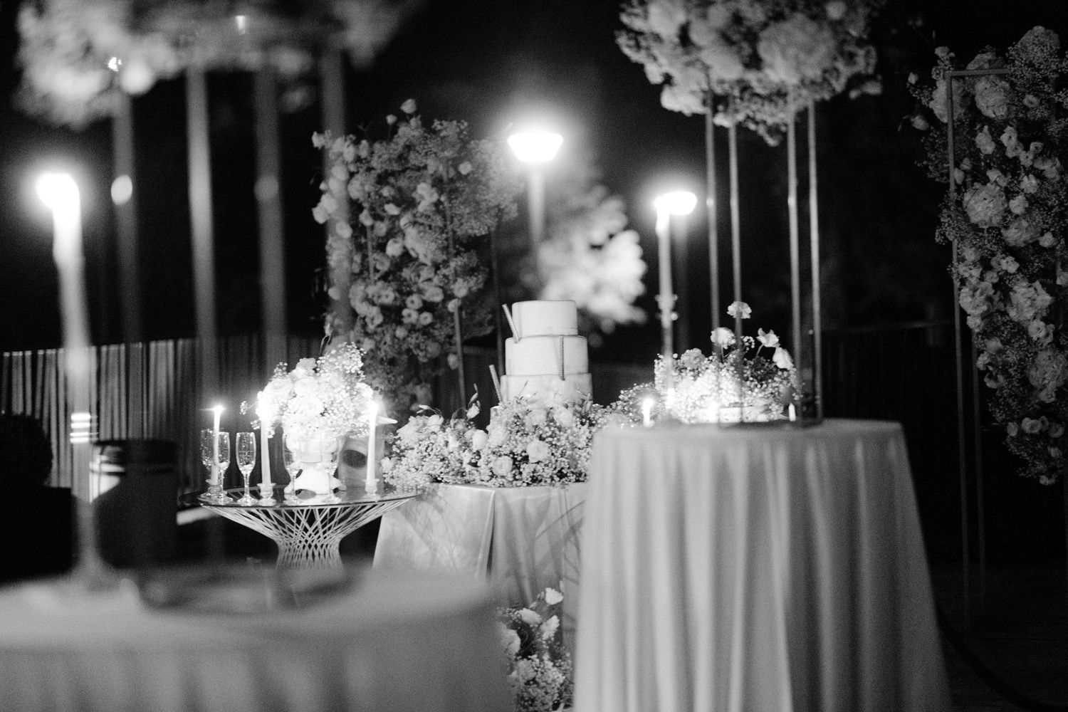Black and white photo of an elegant wedding reception table with floral arrangements and glowing candles at night.