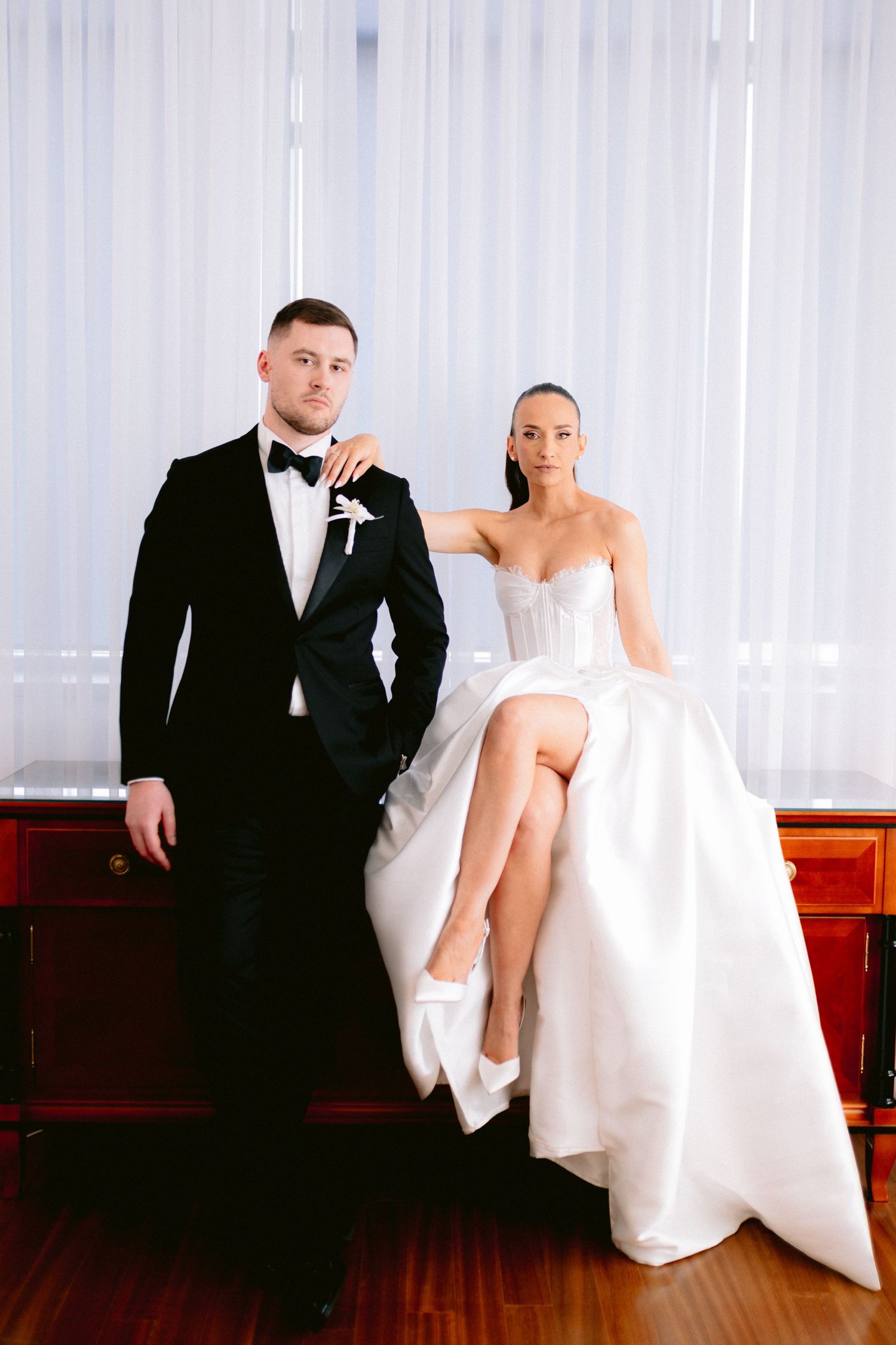 A wedding couple sits together on a red bench in a bright room with white curtains.