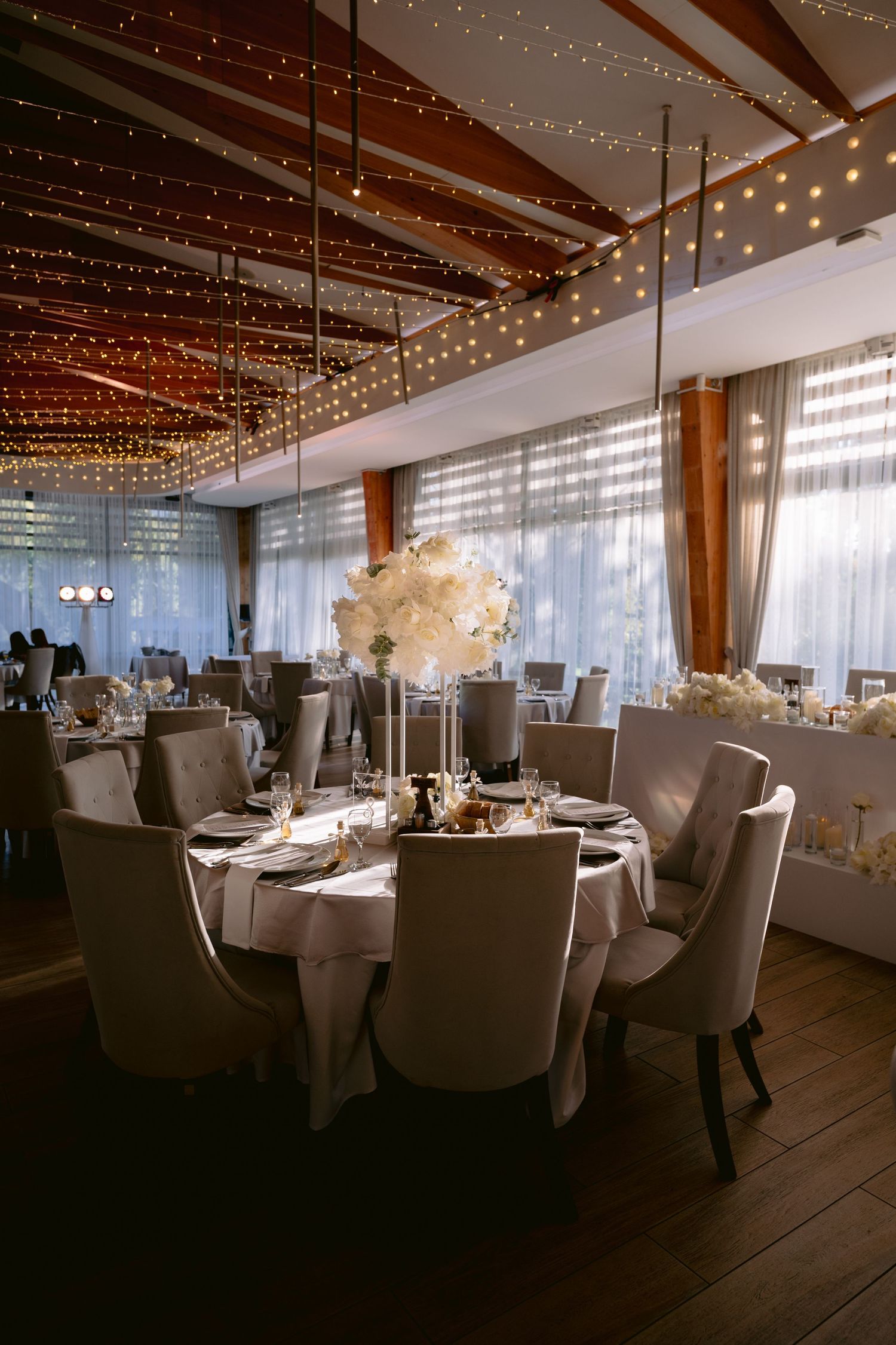 Elegant wedding reception venue with twinkling fairy lights on wooden beams and white-clothed round tables with gray chairs.