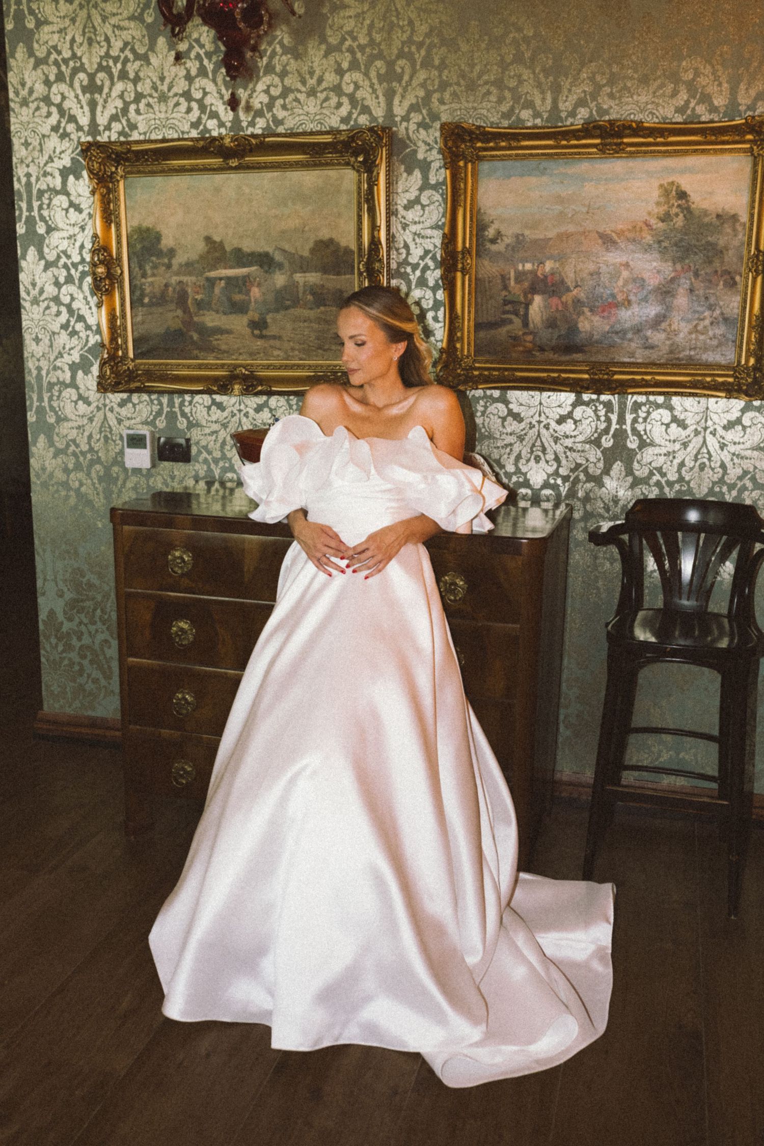 Elegant off-shoulder white satin wedding gown with dramatic train shown in vintage-styled room with landscape paintings.
