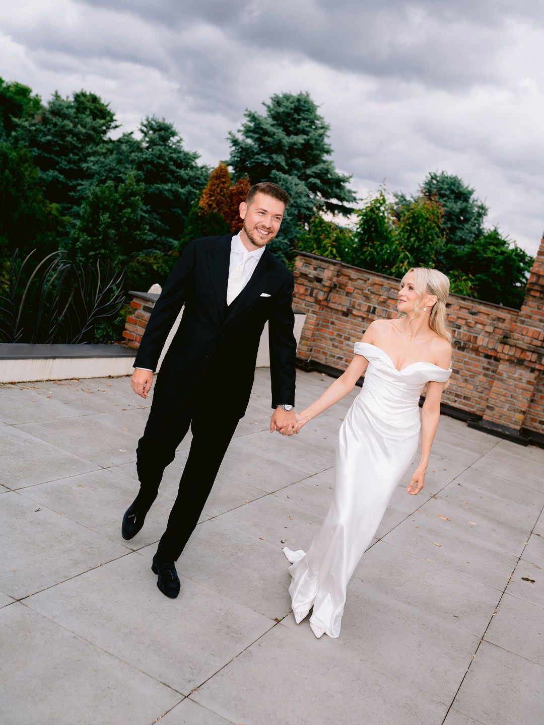 Bride in off-shoulder white satin gown holds hands with groom in black tuxedo walking on stone terrace.
