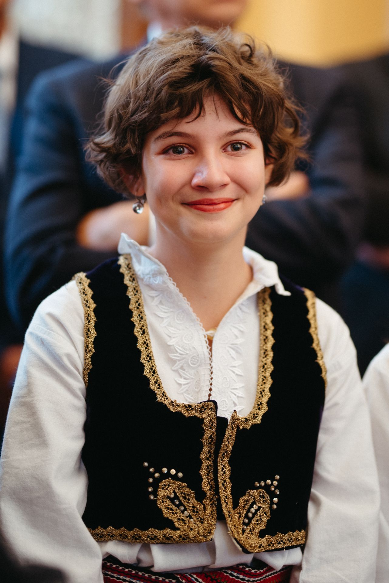 Young woman smiling in traditional Eastern European folk costume with black vest and gold embroidery.