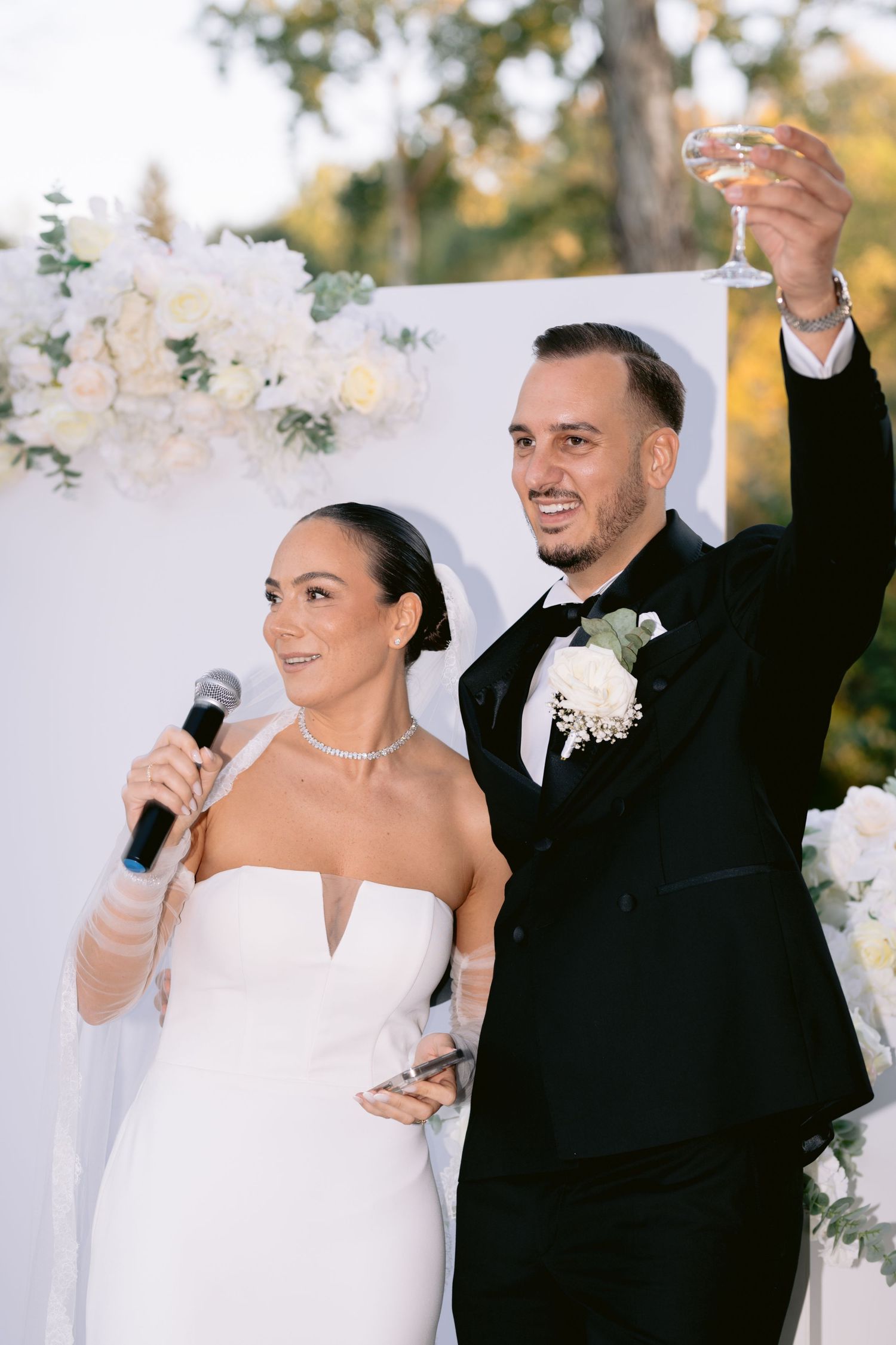 Happy newlyweds celebrate during their outdoor wedding reception with white floral decor in the background.