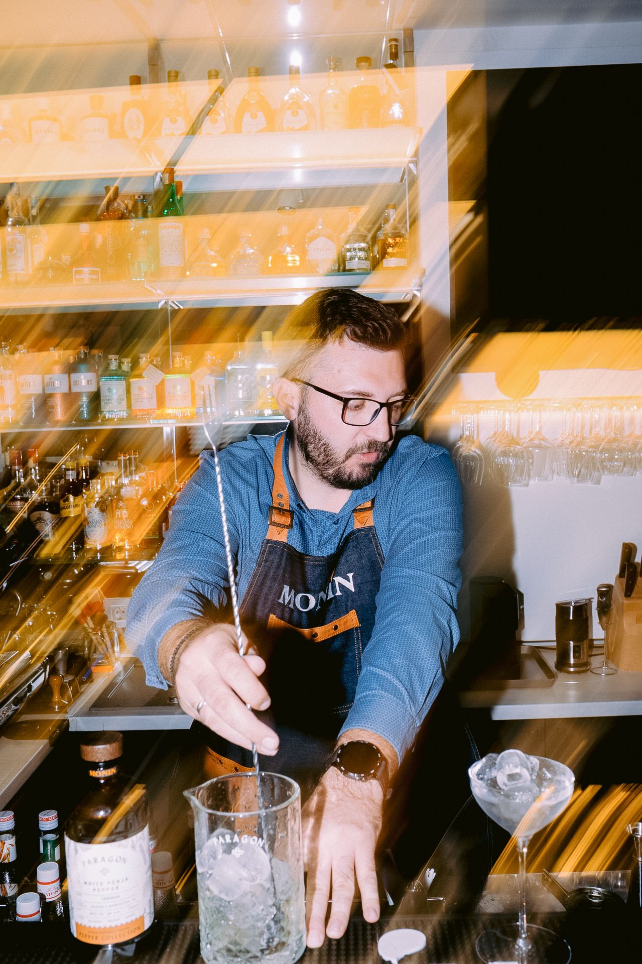 Bartender mixing cocktails behind a well-stocked bar with ambient lighting.