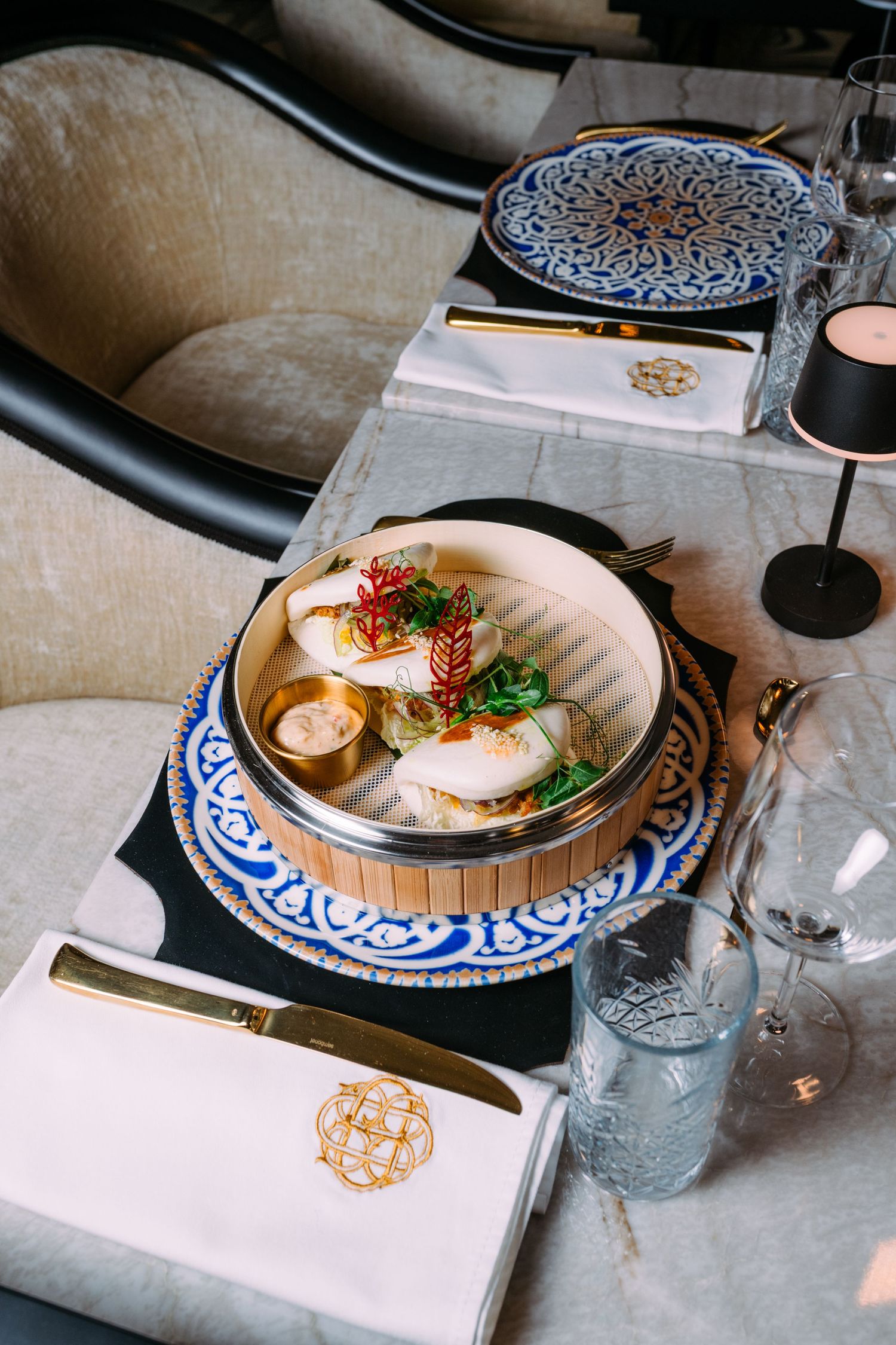 Elegant fine dining table setting with dim sum in a bamboo steamer, blue decorative plates, crystal glasses, and gold cutlery.