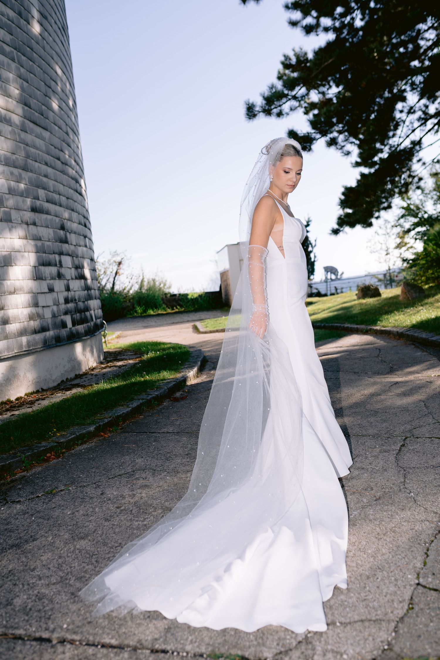 Bride in a white mermaid gown and long veil stands on a path near a stone tower, looking down gracefully.