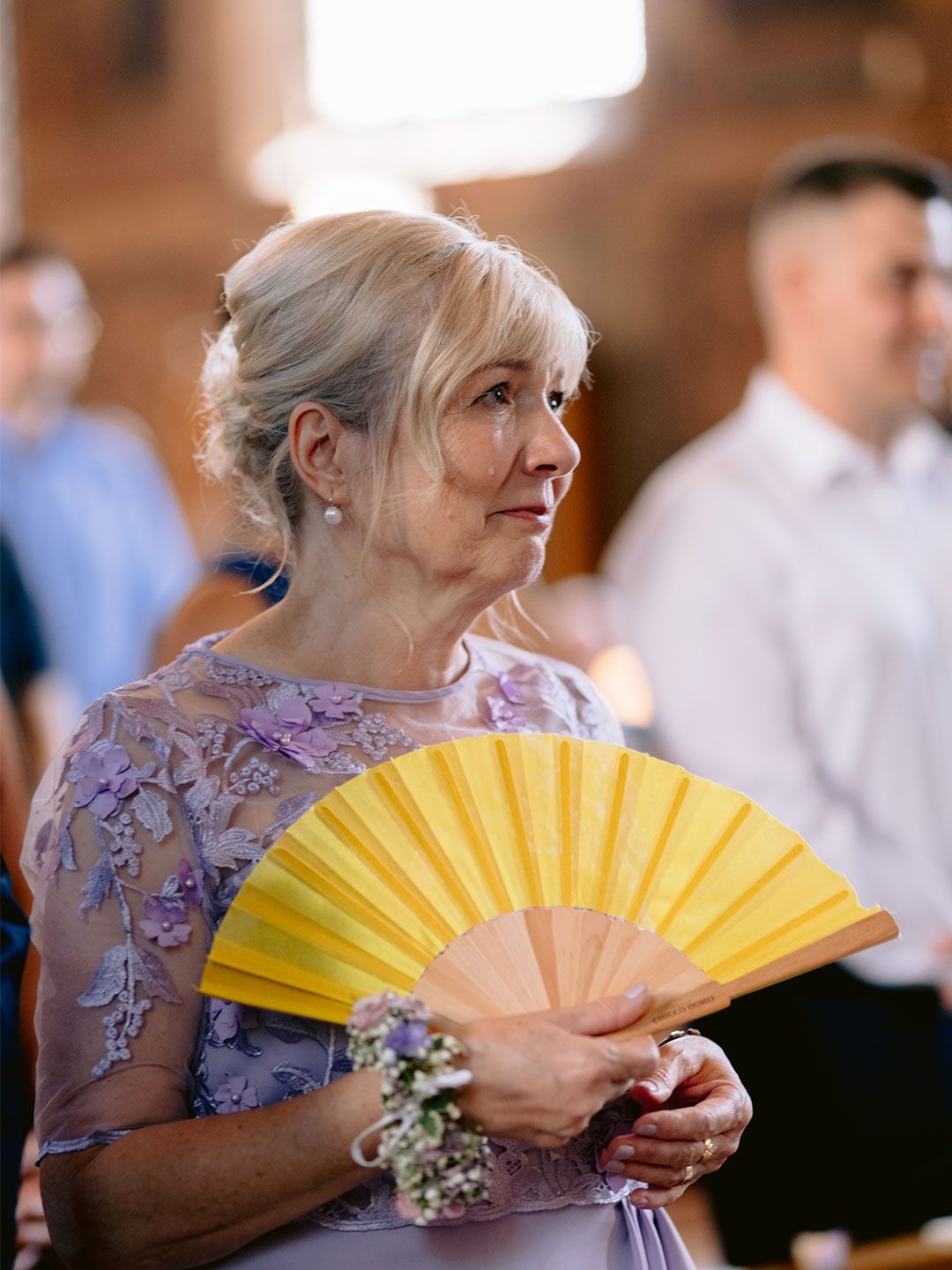 Elegantna starija gospođa u lavanda haljini sa cvjetnim uzorkom i žutom lepezom na ceremoniji vjenčanja.