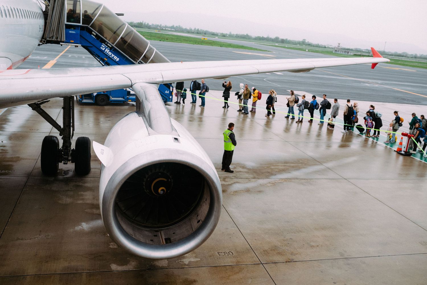 Passengers board a commercial aircraft in the rain while a large jet engine is visible in the foreground.