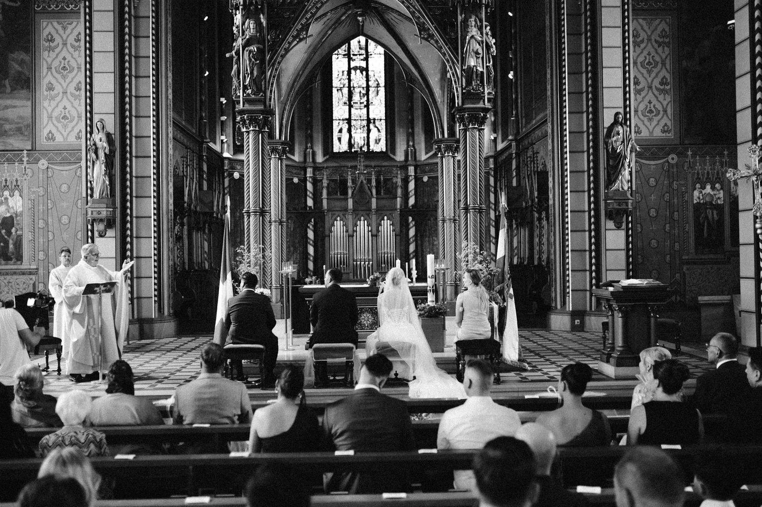 Black and white wedding ceremony photo inside a grand Gothic cathedral with bride, groom, and guests.