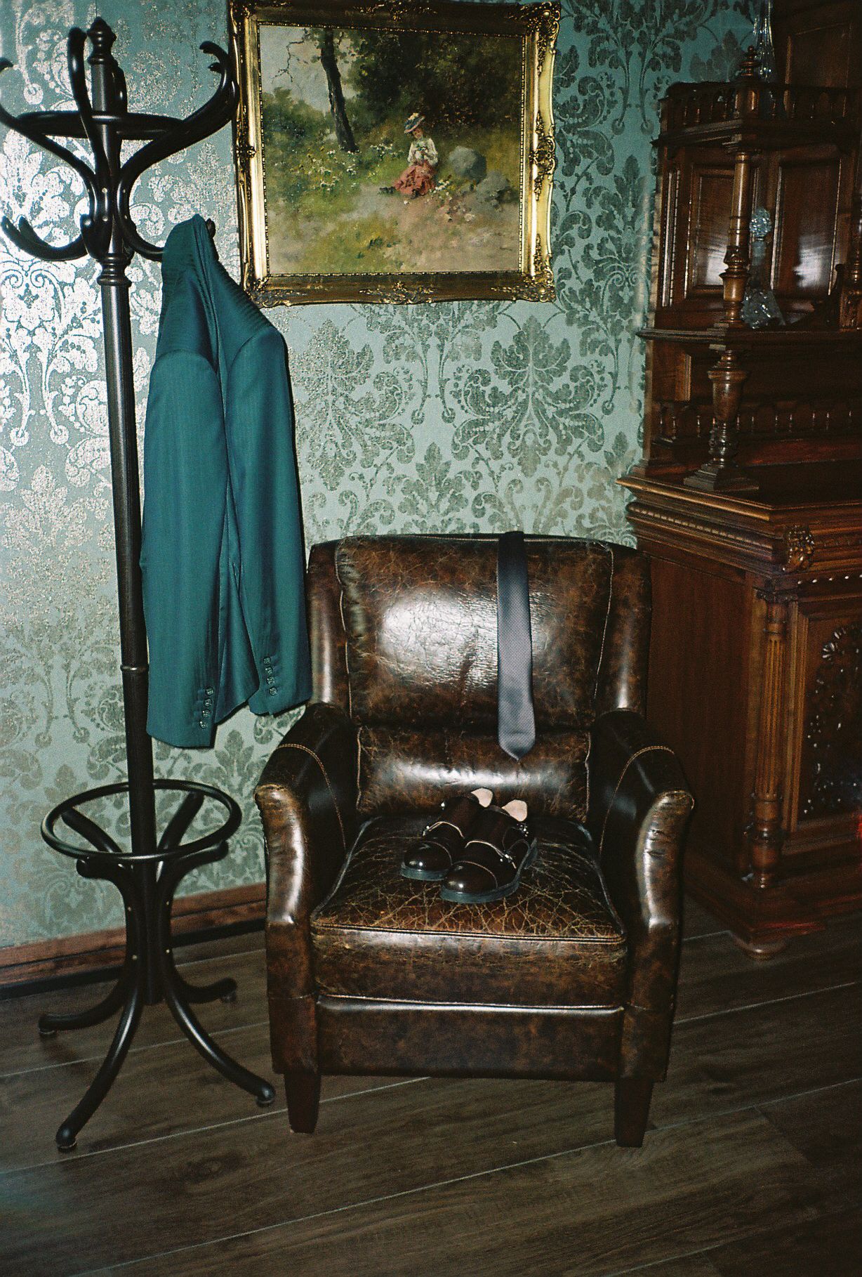 A vintage leather armchair sits against a teal damask wallpaper with an antique coat rack visible in the corner.