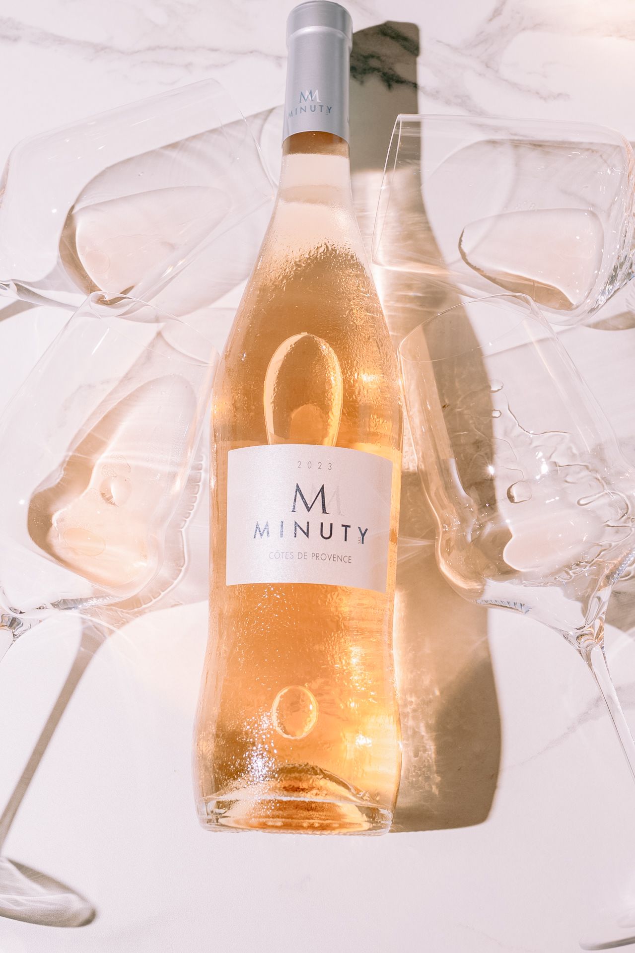 Bottle of rose wine with M branding surrounded by crystal glasses on white surface.