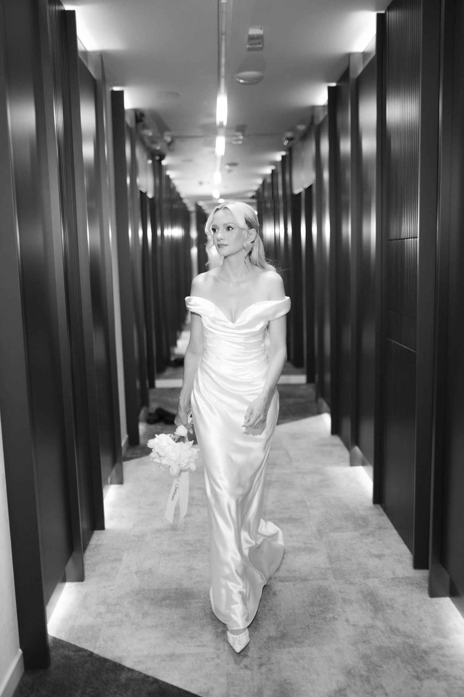 Elegant bride in off-shoulder satin gown walks down a modern hotel corridor in black and white photo.