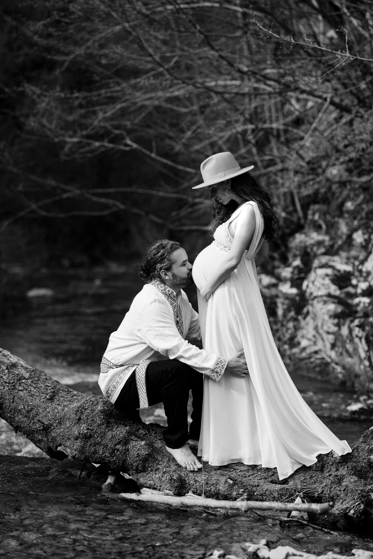 Black and white maternity photo of couple by a creek, man kneeling before pregnant woman in white dress and hat.