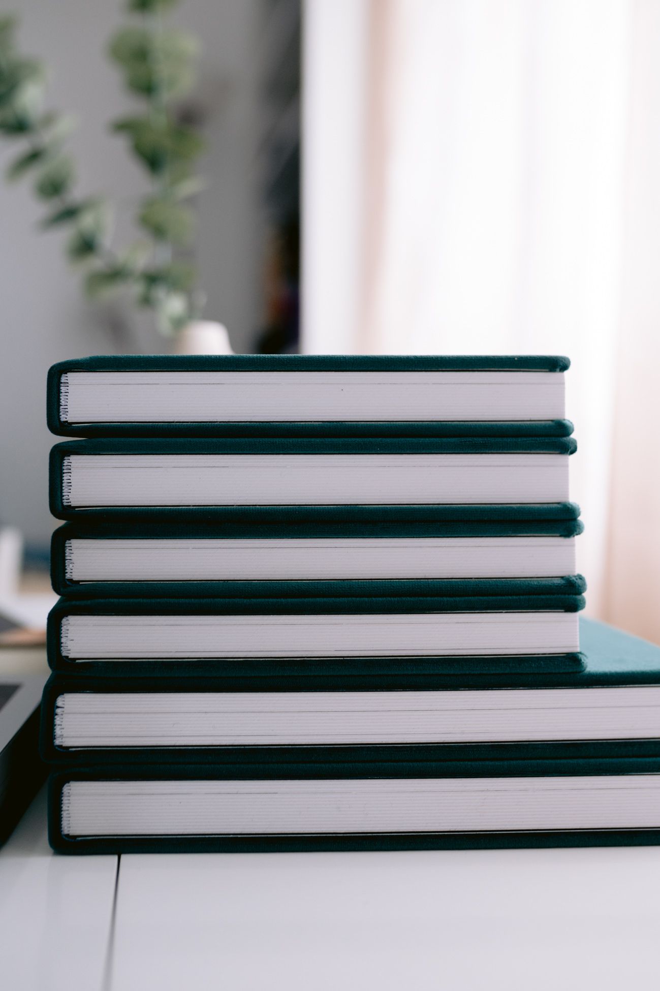 Stack of six dark green hardcover books on a white surface with blurred greenery in the background.