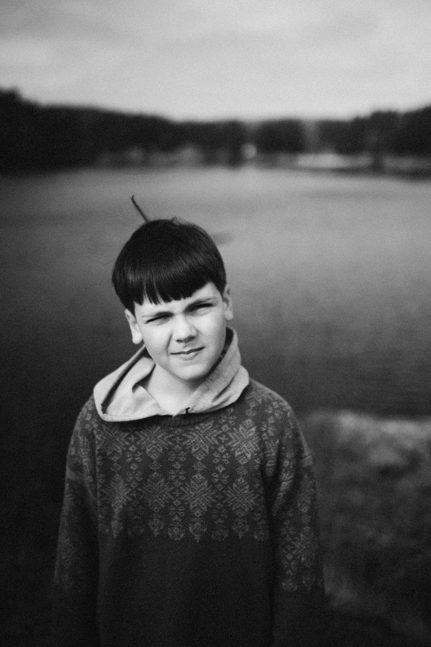 Black and white portrait of a young person in a patterned sweater standing by a lake.