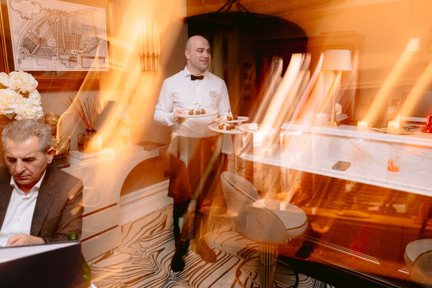 Waiter in bow tie carries dessert plates through elegant restaurant with warm golden light and motion blur effect.