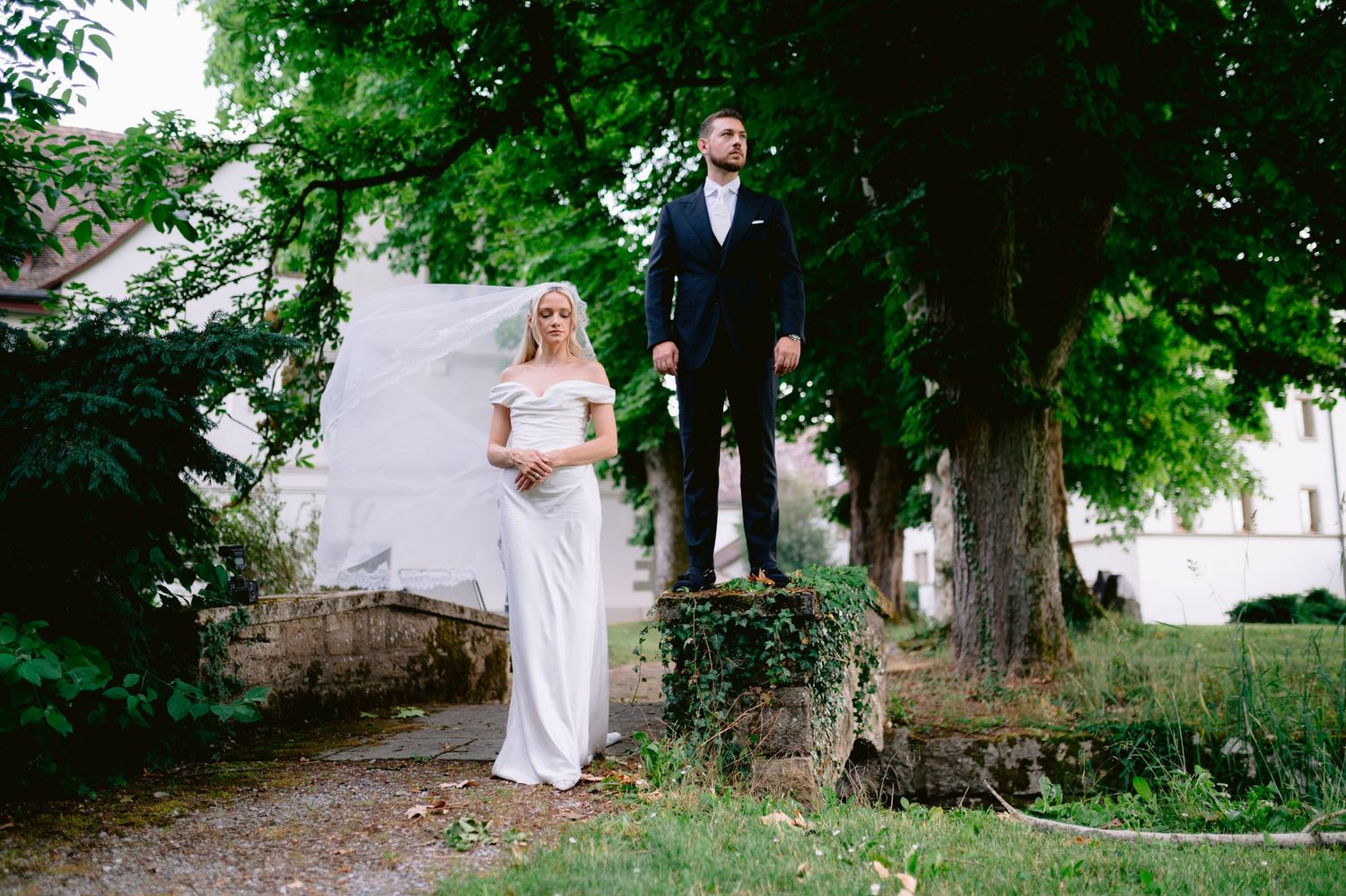 Bride in white gown and groom in navy suit pose outdoors near a tree at a garden wedding venue.