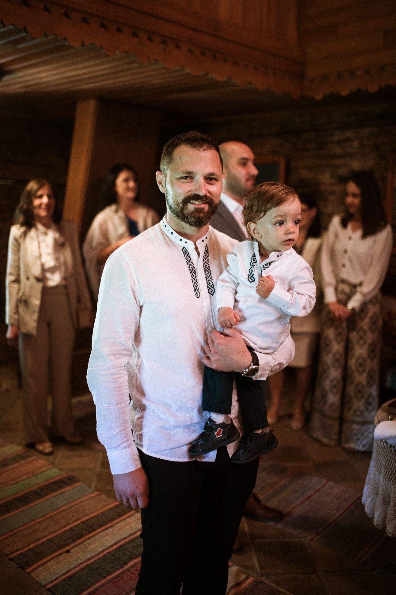 A bearded man in a white embroidered shirt holds a baby at a traditional cultural celebration indoors.