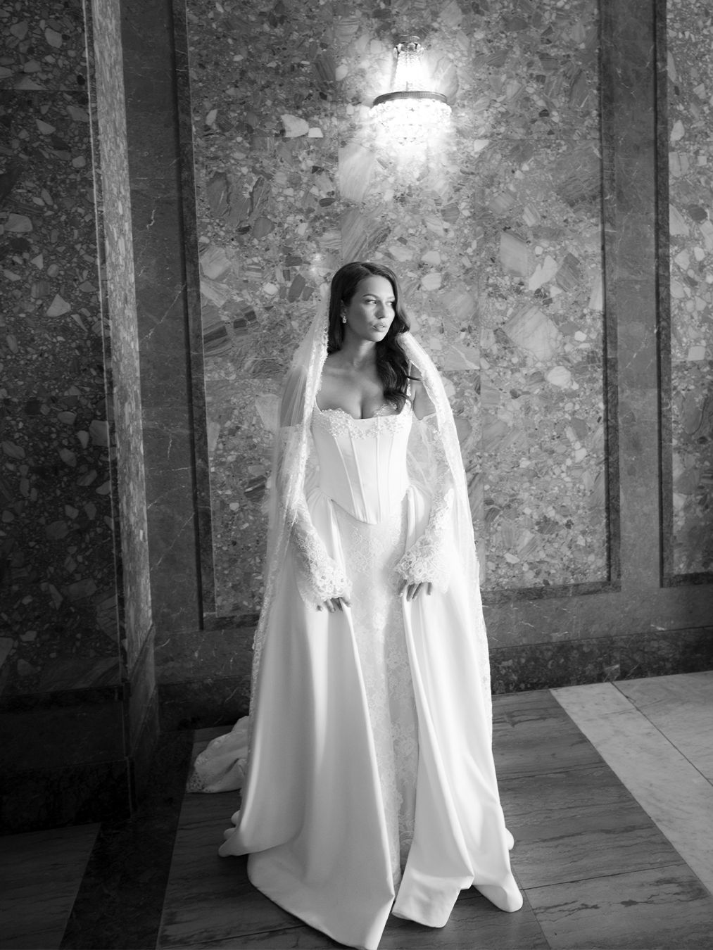 Elegant bride in strapless corset gown with lace sleeves and cathedral veil in marble hall.