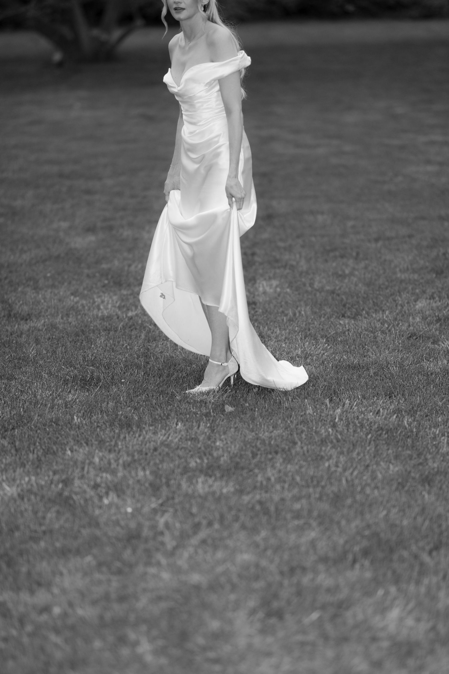 Black and white photo of a bride in an off-shoulder white satin gown walking on green grass.