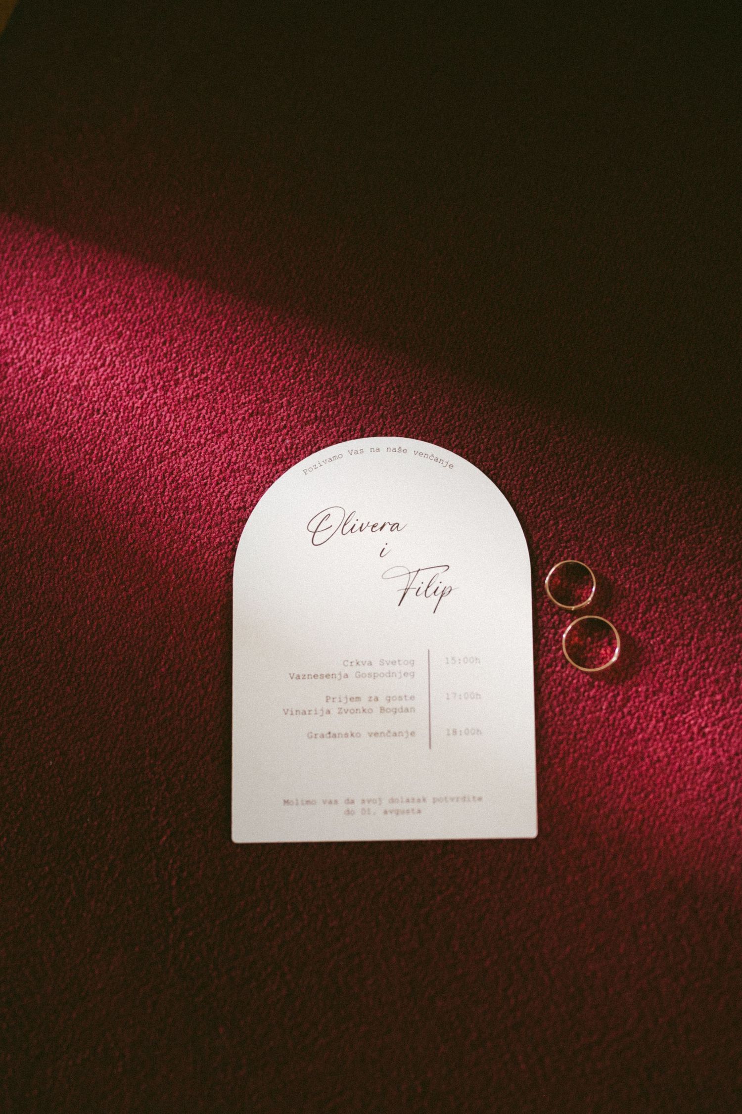 Wedding rings and elegant white invitation cards displayed on a luxurious deep red velvet background.