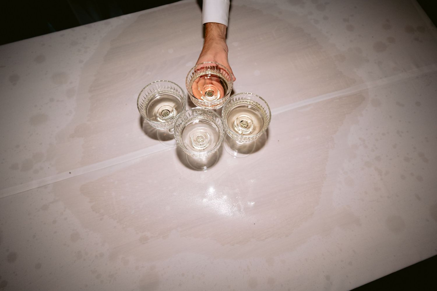 A hand holding a glass of rosé wine above three crystal glasses on a marble table at an elegant event.