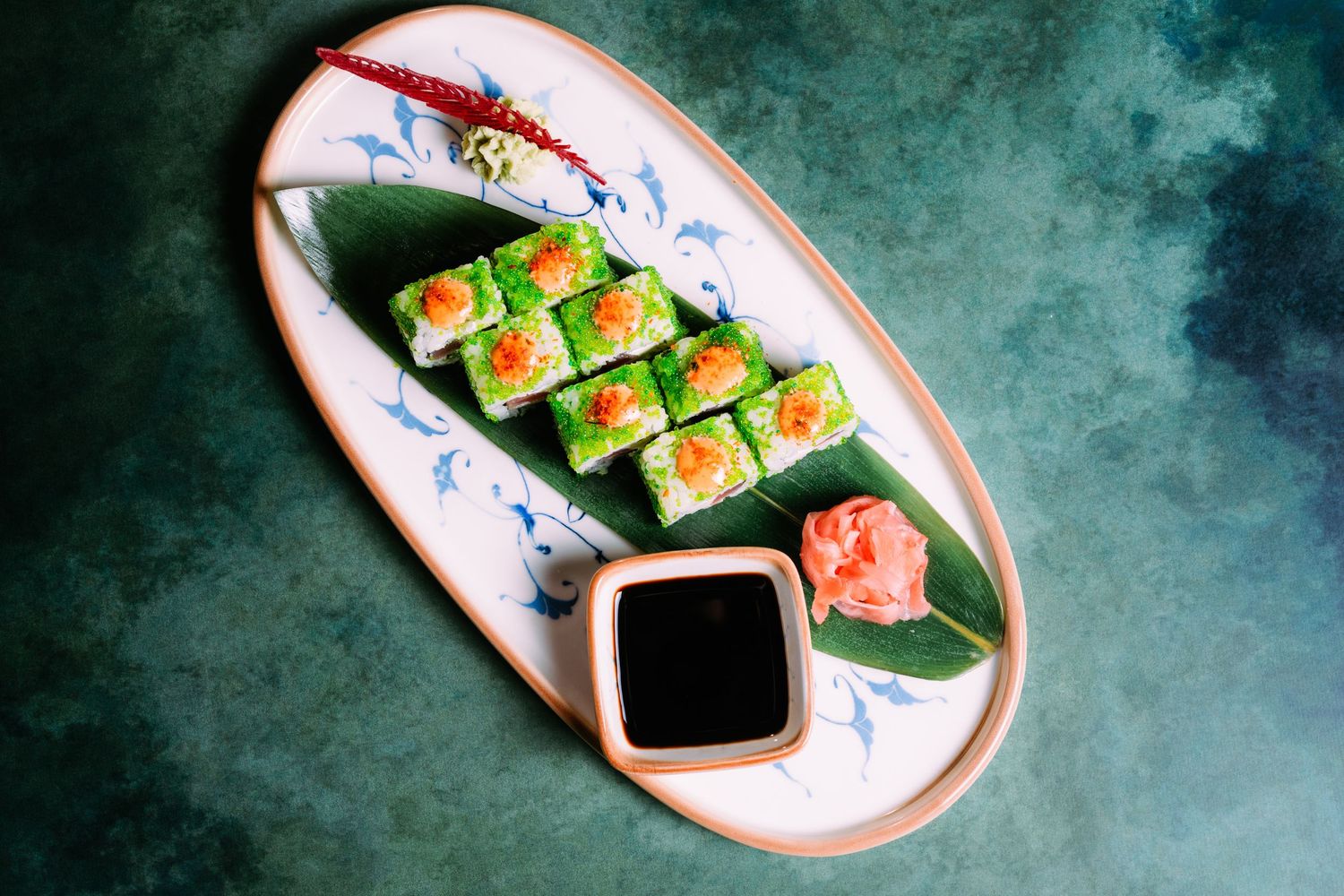 Top-down view of vibrant green sushi rolls topped with orange tobiko served with soy sauce and pickled ginger on a decorative plate.