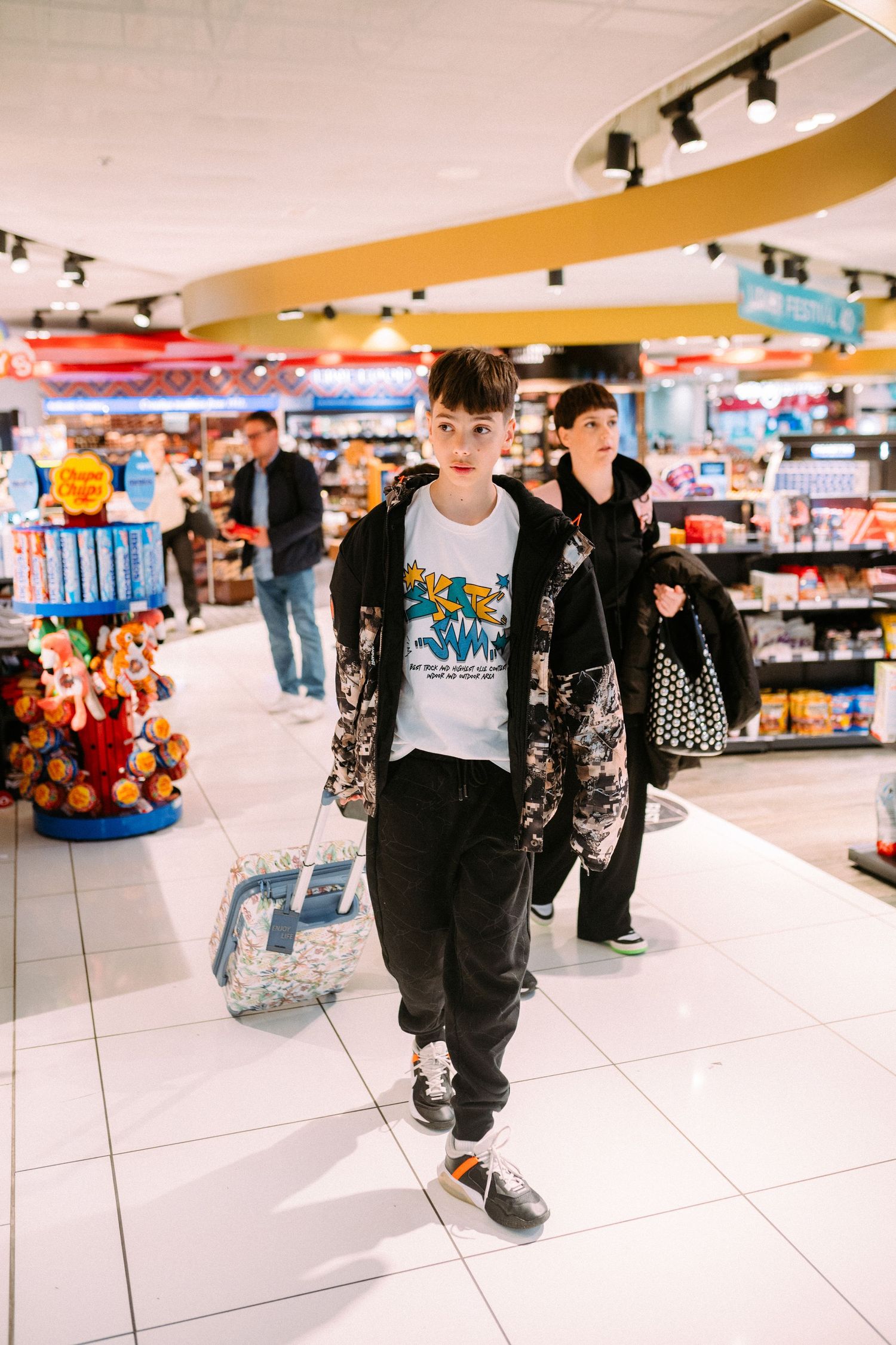 Person wearing black jacket and pants walks through bright retail store with shopping baskets and merchandise displays.