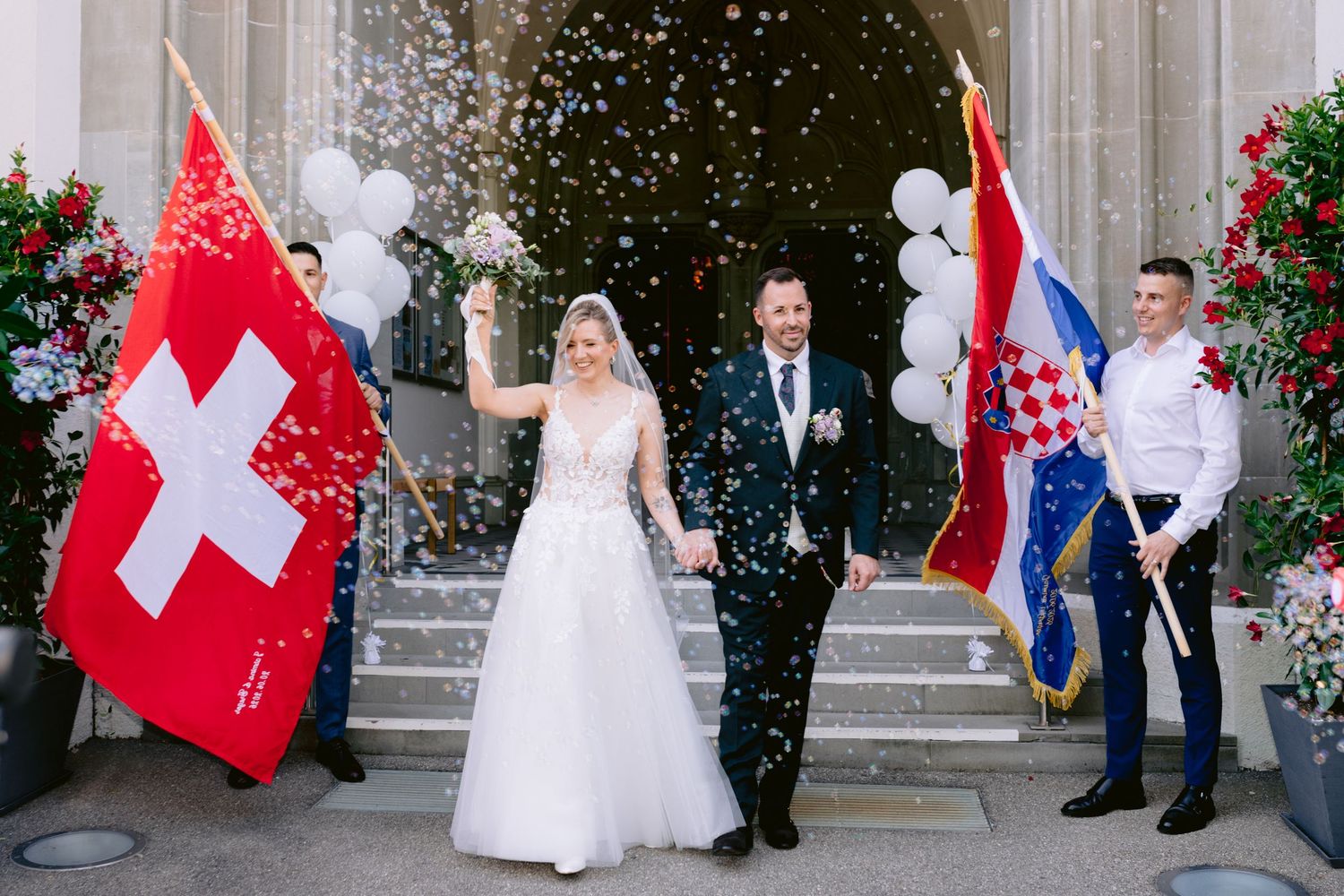 Bride and groom celebrate wedding exit with Swiss and Croatian flags, confetti, and white balloons at church entrance.
