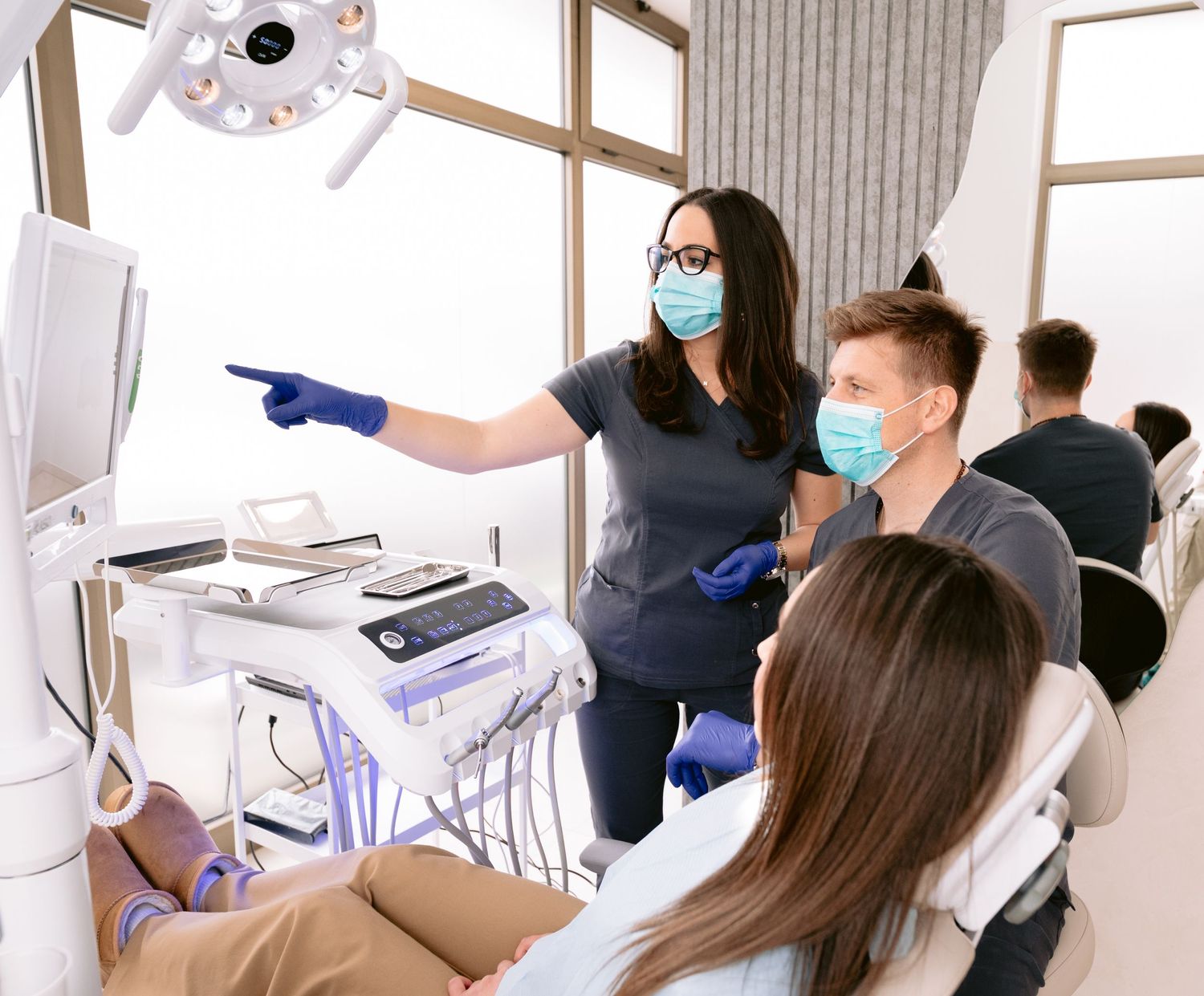 Dentist in blue scrubs and mask shows patient dental X-rays on monitor in modern dental clinic.