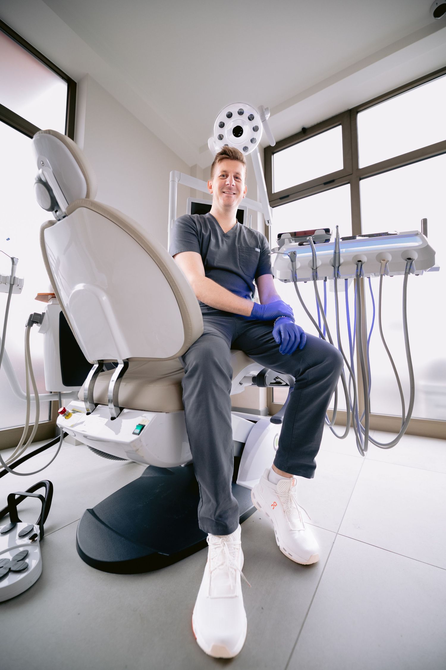 Smiling dentist in scrubs and gloves sitting confidently in a dental chair surrounded by professional dental equipment.