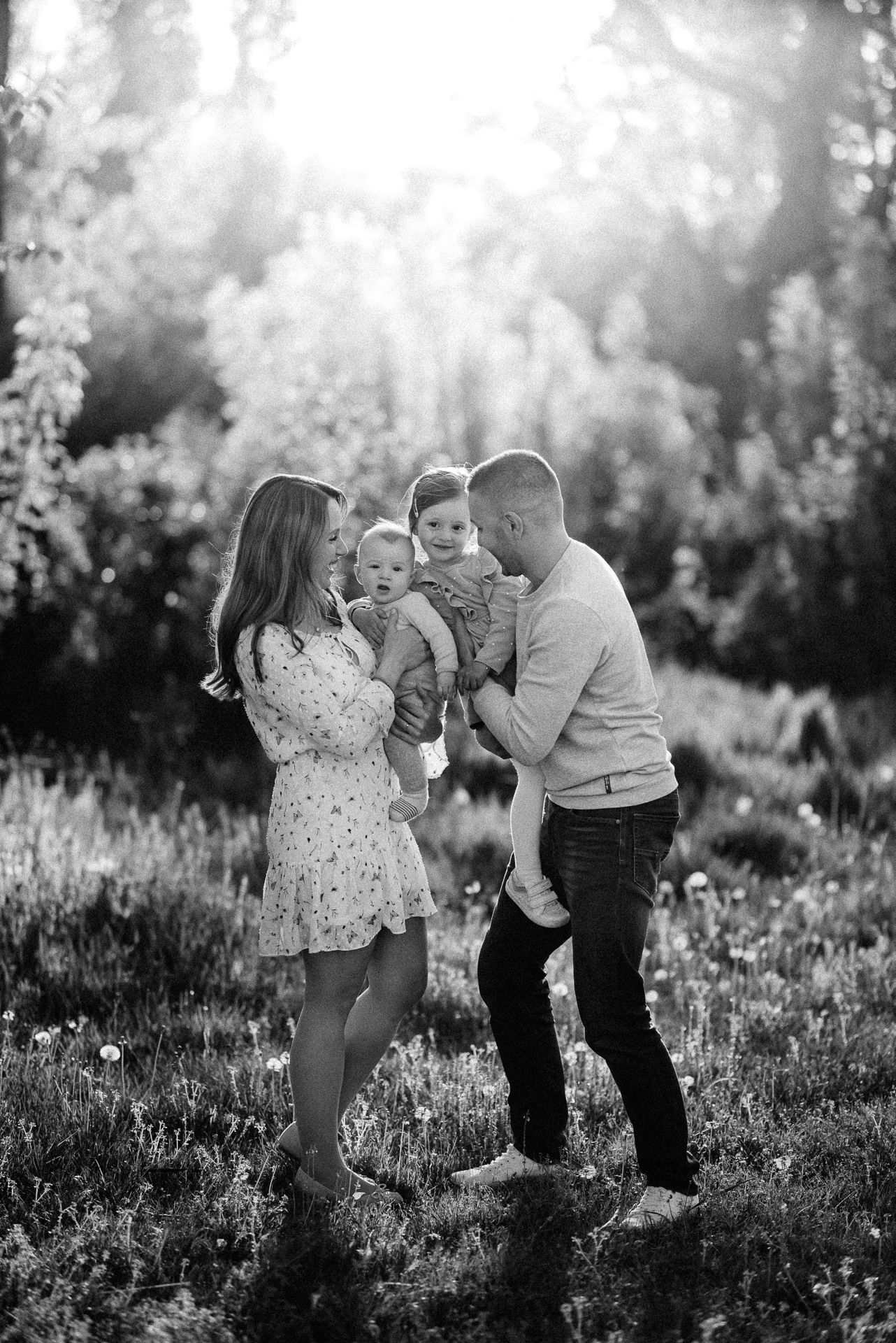 Black and white family photo of parents holding two young children in a sunlit outdoor meadow setting.