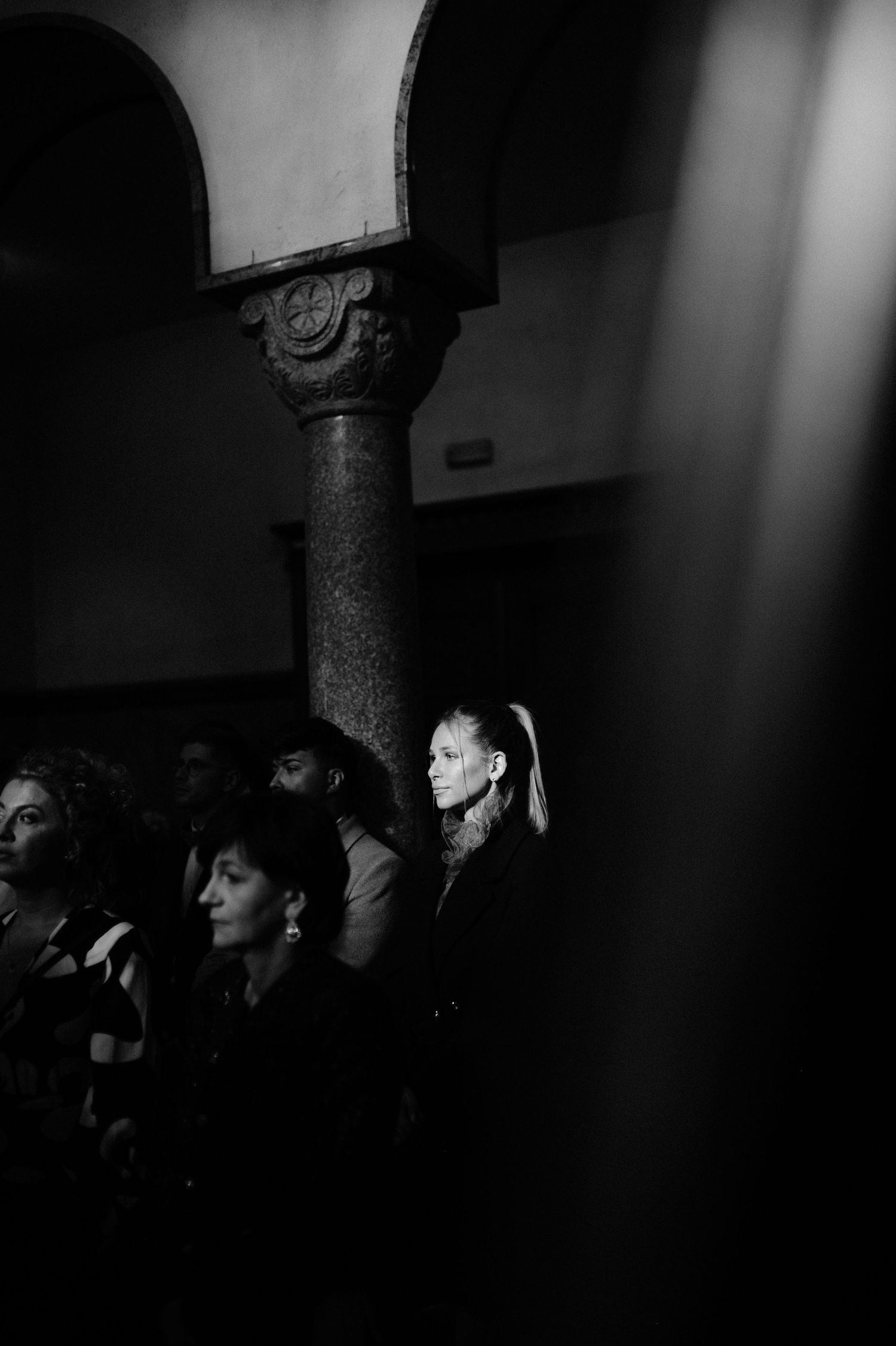 Black and white photo of elegant audience members in a historic arched hall, woman with ponytail in sharp focus.