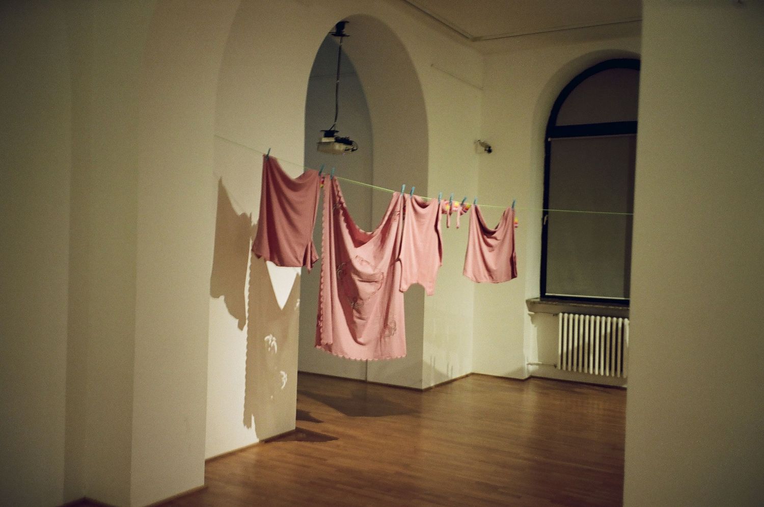 Pink fabric garments hang suspended in an empty gallery space with white walls and arched doorways.