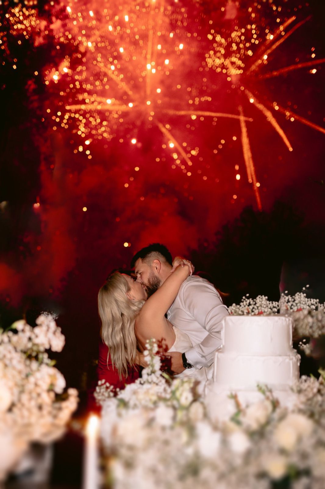 Romantic wedding kiss moment under spectacular red fireworks display with white floral decorations in foreground.
