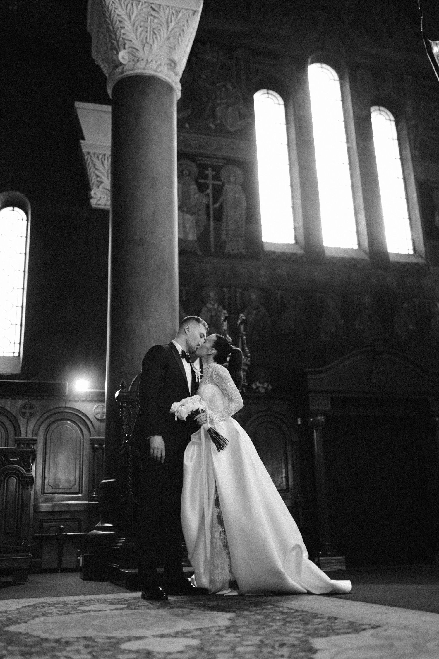 Crno-bijela fotografija mladenaca u zagrljaju u crkvi Svete Trojice u Banja Luci sa zracima svjetla kroz prozor.