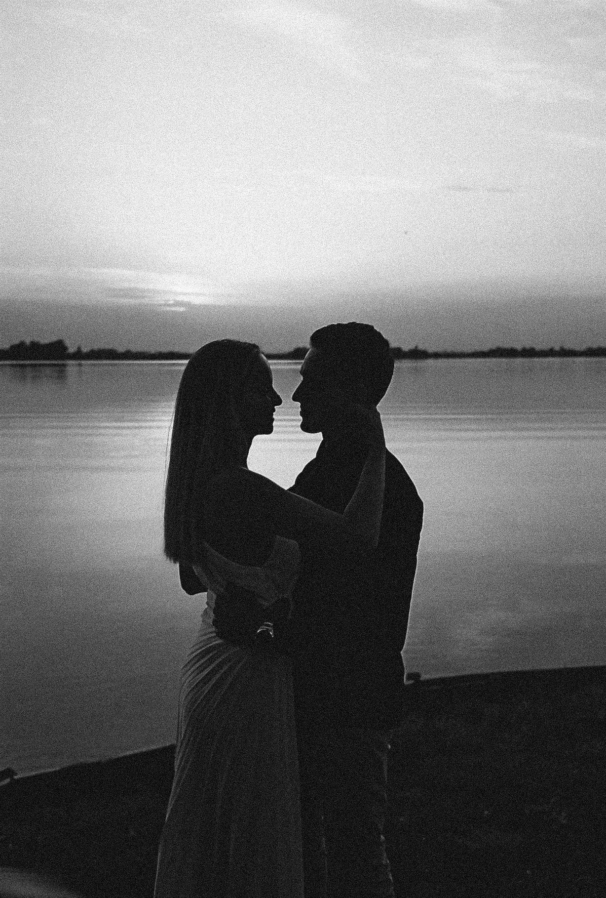 A romantic silhouette of a couple embracing by a serene lake at sunset in black and white photography.