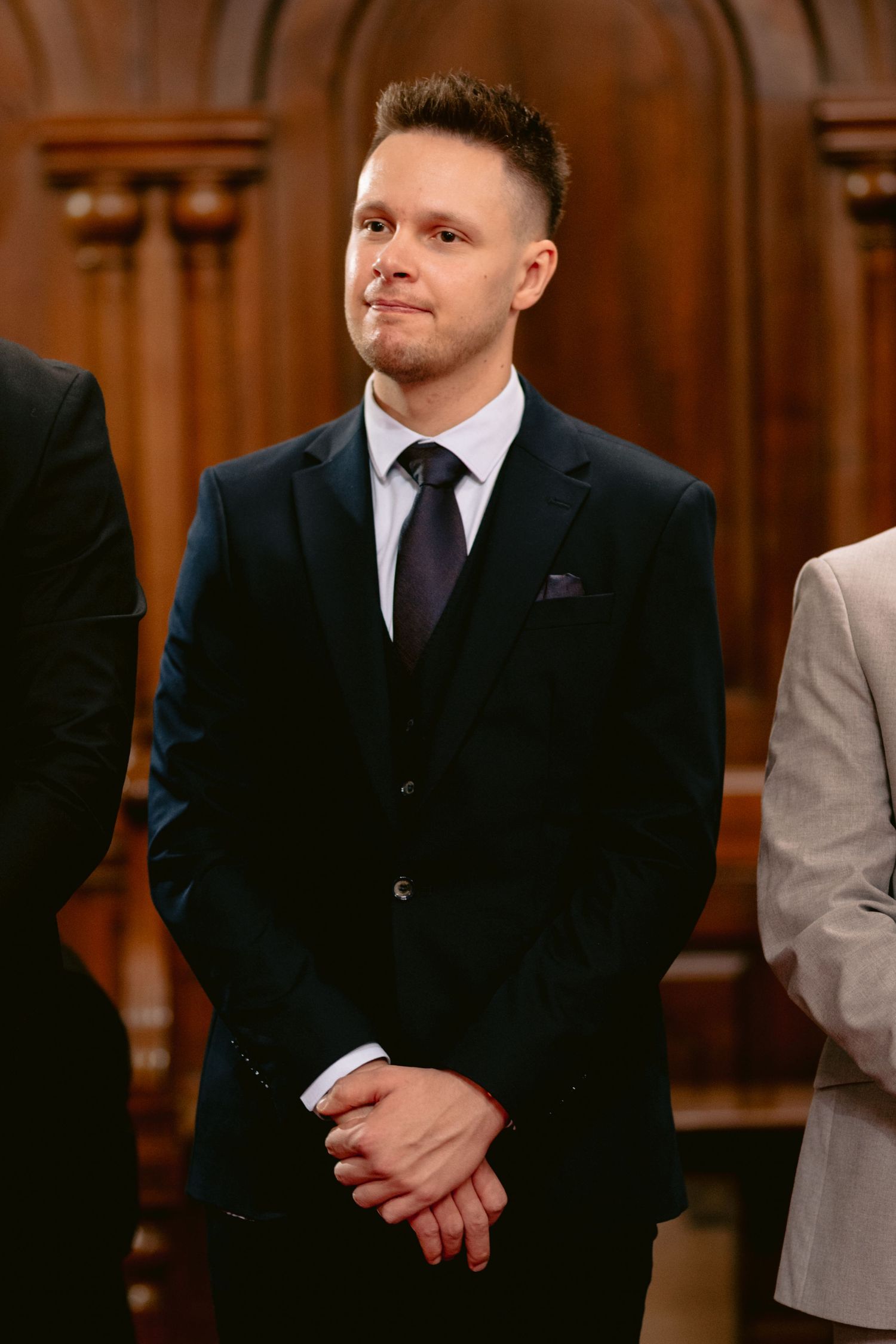 A person wearing a black suit and tie stands in a formal setting with warm wooden paneling in the background.