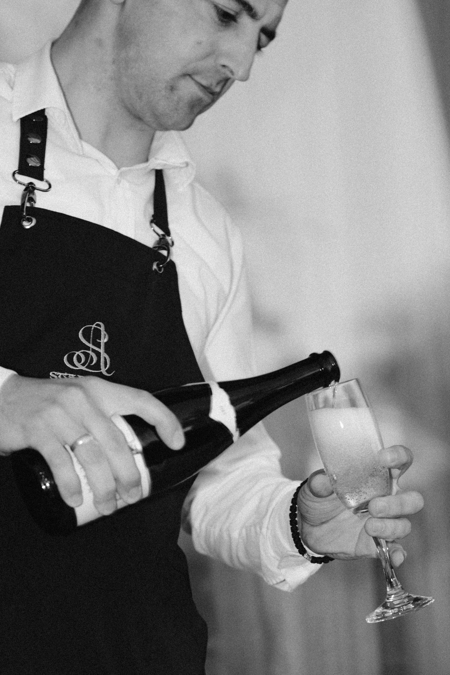 A server wearing a black apron pours champagne into a delicate glass in this elegant black and white photograph.