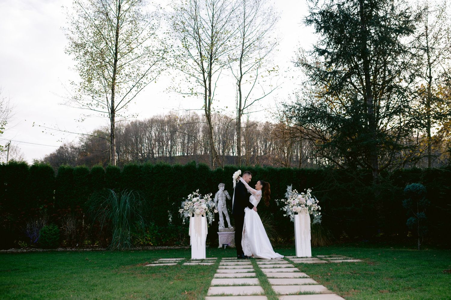 Wedding party walks along a stone pathway through lush gardens at an elegant outdoor ceremony venue.