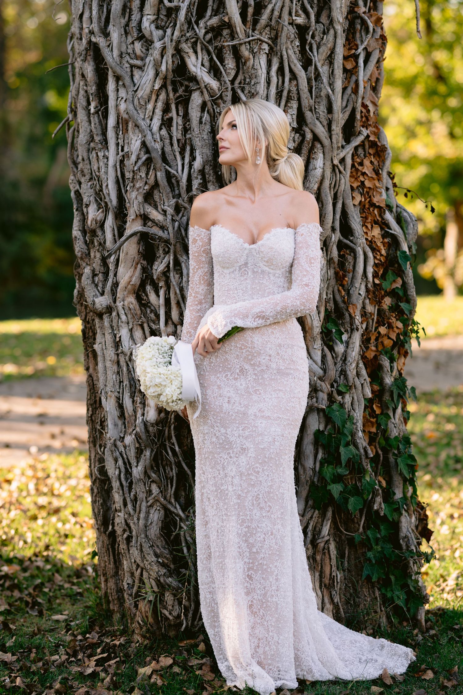 Bride in form-fitting lace gown poses against textured tree trunk in autumn setting.