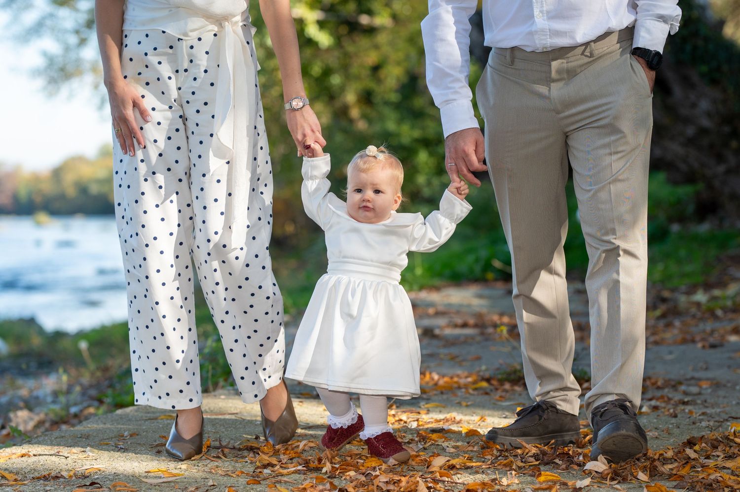 A toddler in a white dress holds the hands of two parents while walking on a leaf-covered path beside a calm lake in autumn.