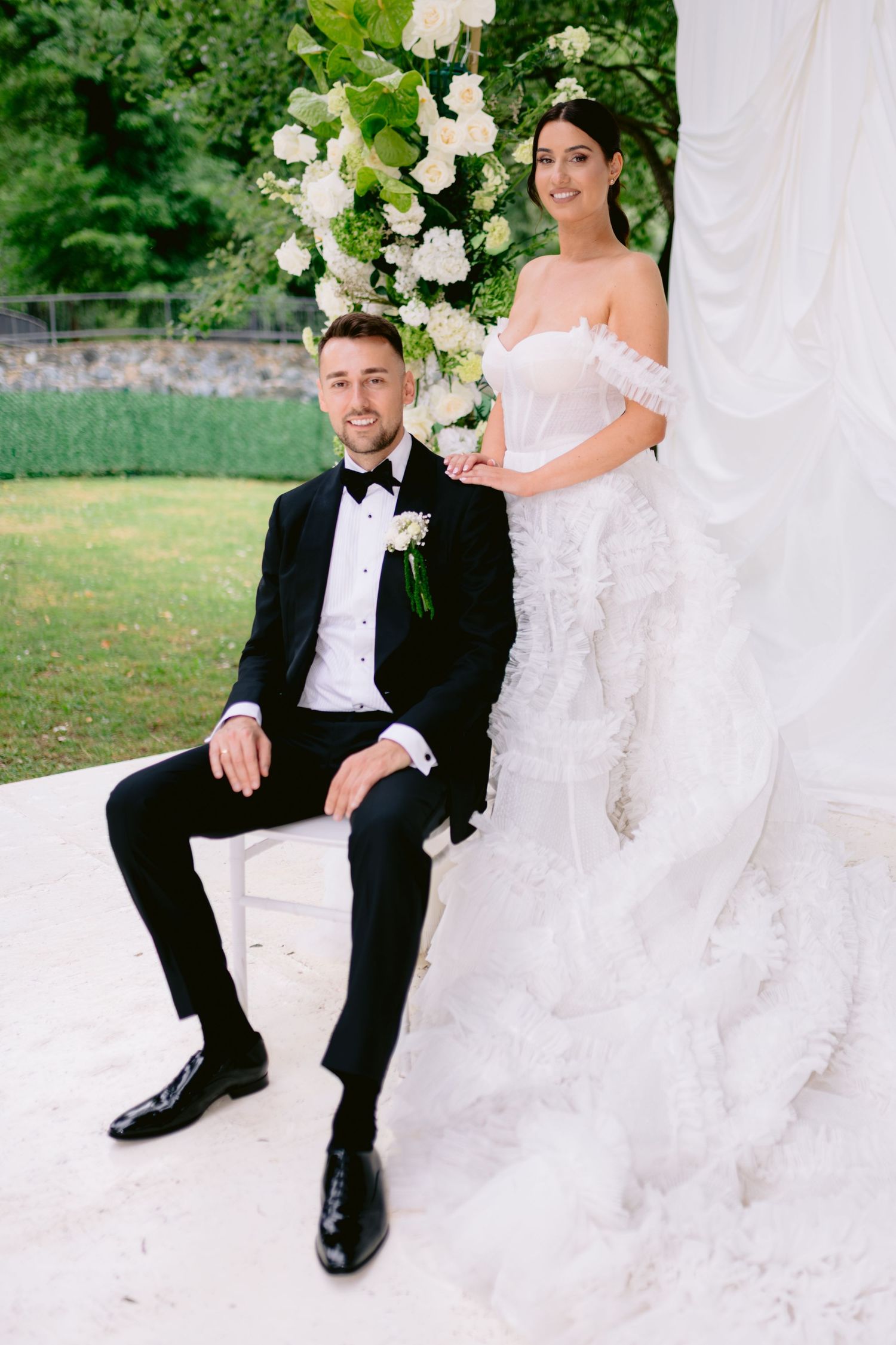 Elegant bride in strapless ruffle gown stands beside seated groom in tuxedo at floral wedding altar.